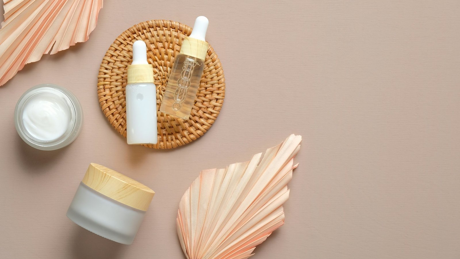 Skincare bottles and jars styled with dried palm leaves on a neutral beige background